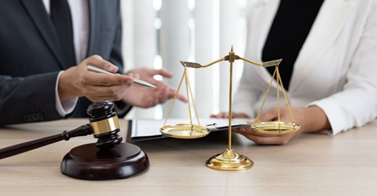 a woman and a man wearing business suits sitting behind a desk, on the desk there is a gavel and legal scales