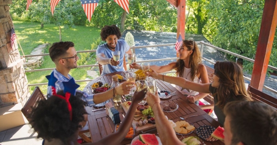 group of people sitting at a covered patio table clinking glasses, there are American flags and other Fourth of July decor hanging from the ceiling