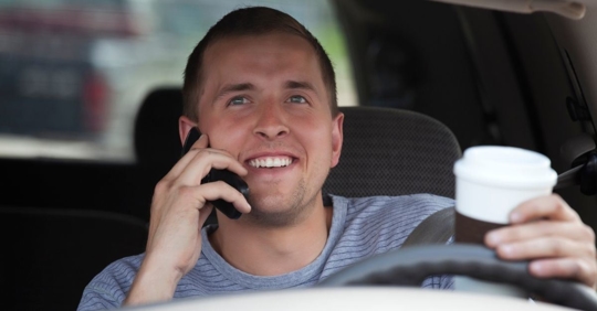 person acting out the three types of distracted driving in a vehicle while talking on a cellphone, looking away from the road, and holding a to-go coffee cup