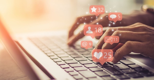 hands typing on a laptop computer, social media "like," "comment," and "message" icons are floating around